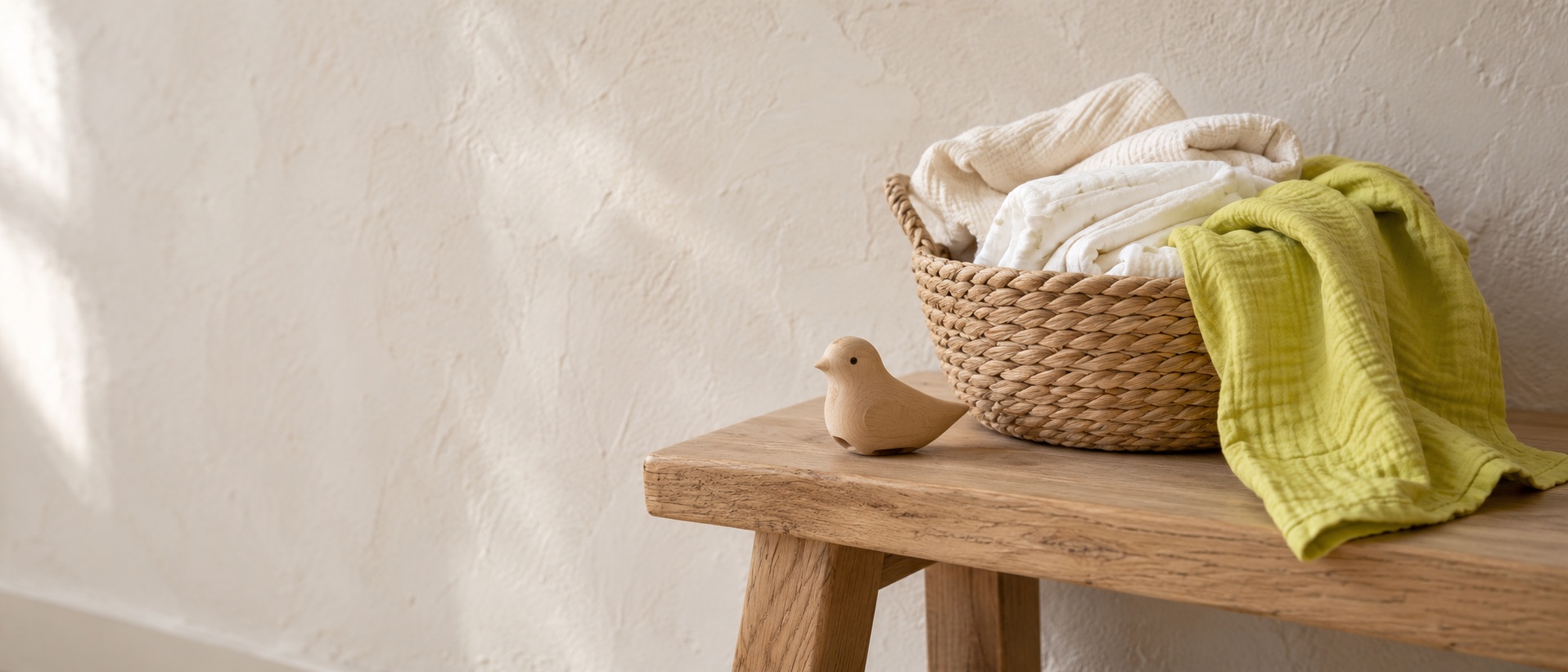 Korb mit hellen Babysachen, grünem Musselintuch und kleinem Holzvogel auf einer Holzbank